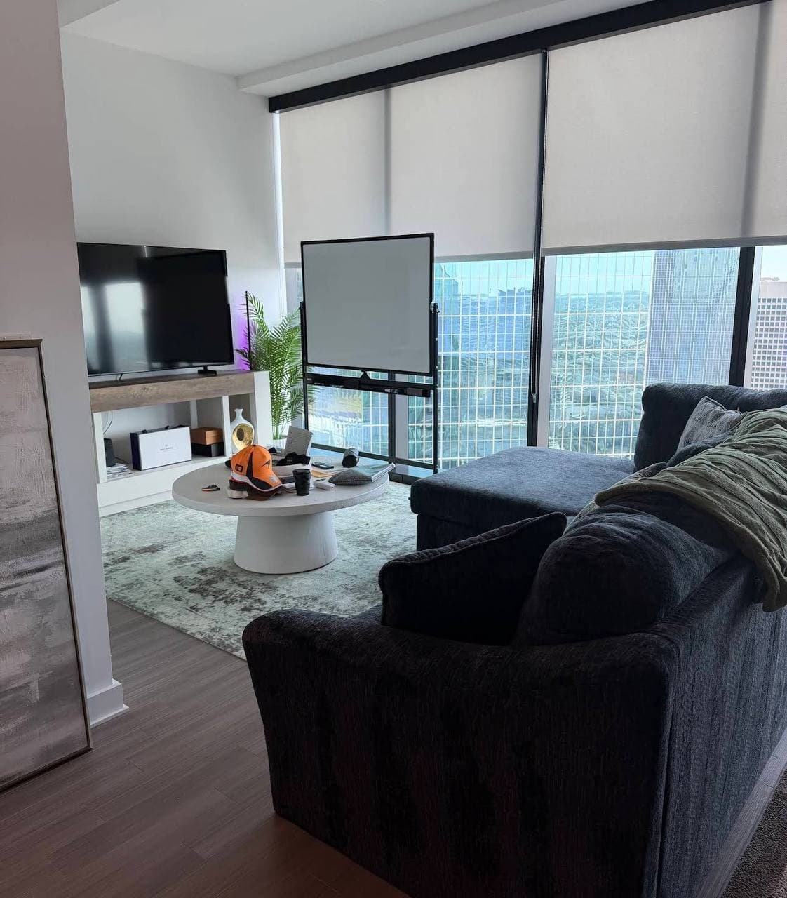 A living room with dark seating, a TV, and a freestanding whiteboard in front of tall windows.